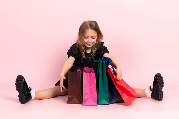 Kinder winkelen. Charmant klein meisje in zwarte stijlvolle zomerjurk en lichte make-up zit op de vloer op zoek in boodschappentassen bij haar nieuwe aankopen. Geïsoleerd op roze achtergrond. - Foto, afbeelding