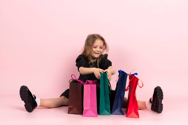 Nakupování dětí. Okouzlující holčička v černých stylových letních šatech a lehký make-up sedí na podlaze a dívá se v nákupních taškách na své nové nákupy. Izolované na růžovém pozadí. - Fotografie, Obrázek