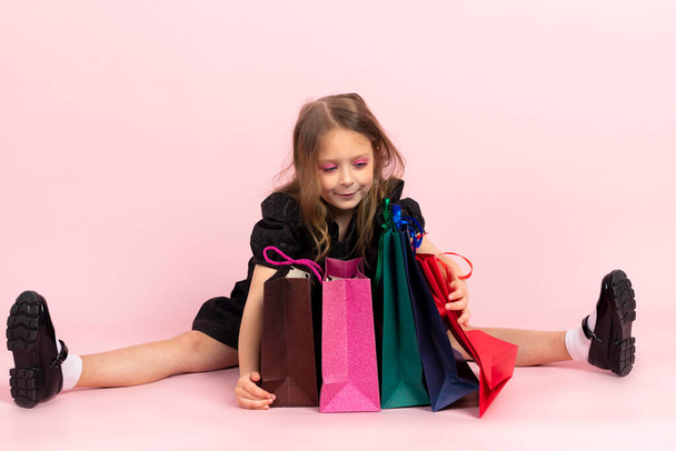 Child shopping. Charming little girl in black stylish summer dress and slight makeup sits on the floor looking in shopping bags at her new purchases. Isolated on pink background. - Φωτογραφία, εικόνα