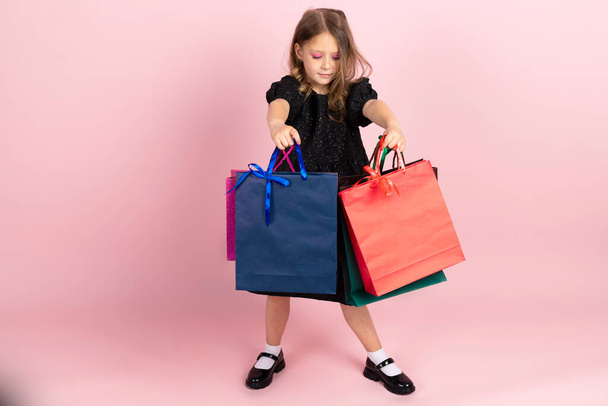 Kinder winkelen. Charmant klein meisje in zwarte stijlvolle zomerjurk en lichte roze make-up, poseren met kleurrijke boodschappentassen. Geïsoleerd op roze achtergrond. - Foto, afbeelding