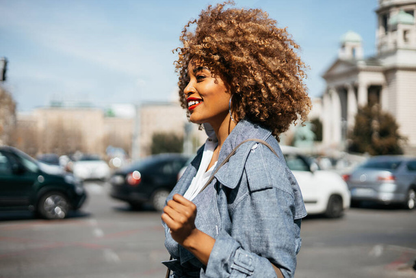 Beautiful and confident black woman walking down the street. She is happy and smiled. Bright sunny day. Side view. - Φωτογραφία, εικόνα