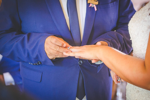Tento krásný obraz zachycuje intimní okamžik, kdy si pár vyměňuje snubní prsteny na skutečné svatbě. Fotografie zblízka zachycuje ruce páru, ukazuje jejich propletené prsty a snubní prsteny na prstech. Obrázek - Fotografie, Obrázek