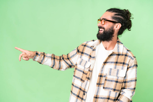 Young man with beard over isolated chroma key background pointing finger to the side and presenting a product - Foto, imagen