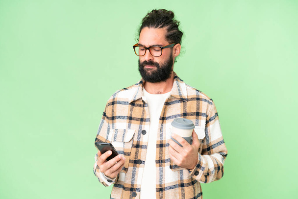Jeune homme avec barbe sur fond isolé chroma clé tenant café à emporter et un mobile - Photo, image