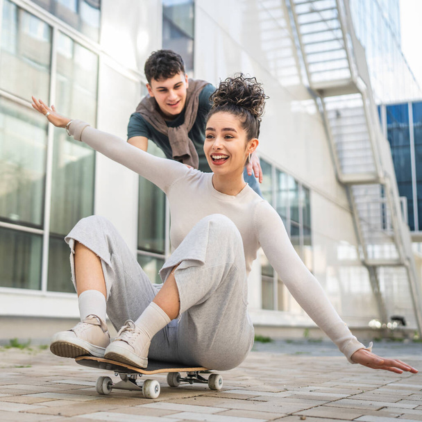 Young man and woman teenager couple caucasian brother and sister have fun on skateboard pushing to ride by the modern building in day happy smile real people family love concept copy space - Фото, зображення