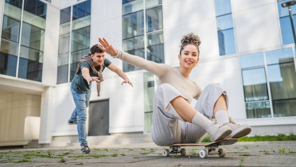 Young man and woman teenager couple caucasian brother and sister have fun on skateboard pushing to ride by the modern building in day happy smile real people family love concept copy space - Photo, Image
