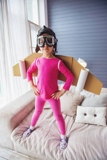 Cute Little Girl ubrany jak pilot z zabawkami skrzydła patrzy na kamerę podczas gry w domu - Zdjęcie, obraz
