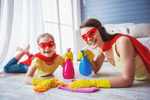 Mooie moeder en haar schattige kleine dochter gekleed als superhelden houden sprayer, kijken naar de camera en glimlachen tijdens het schoonmaken van het huis - Foto, afbeelding