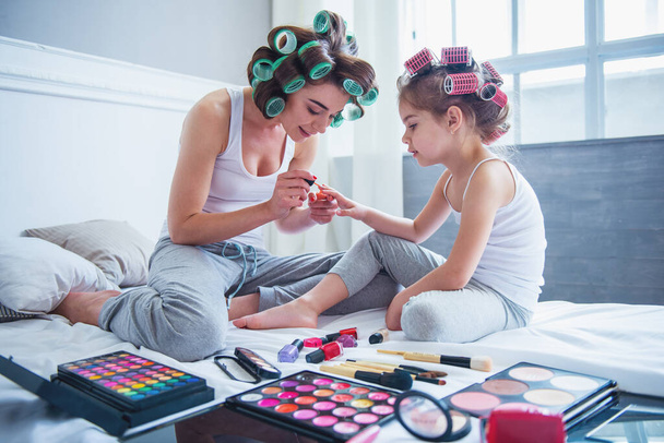 Bela jovem mãe e sua filha com encrespadores de cabelo estão sentados na cama em casa. Mãe está fazendo sua filha manicure
 - Foto, Imagem