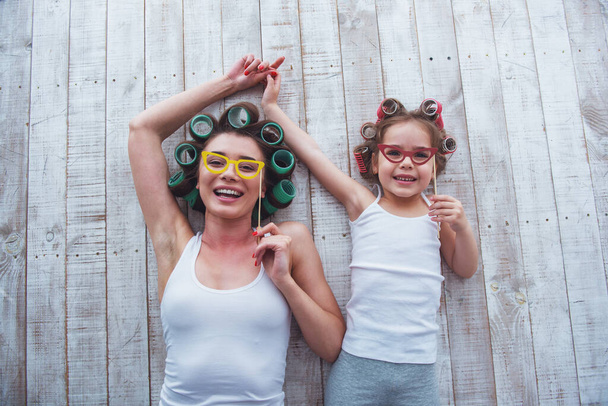 Κάτοψη του νεαρή μητέρα και την κόρη της με τα σίδερα μαλλιών στο κεφάλι τους κρατώντας γυαλιά εγγράφου σε ραβδί και χαμογελαστός, κείνται επί του δαπέδου στο σπίτι - Φωτογραφία, εικόνα