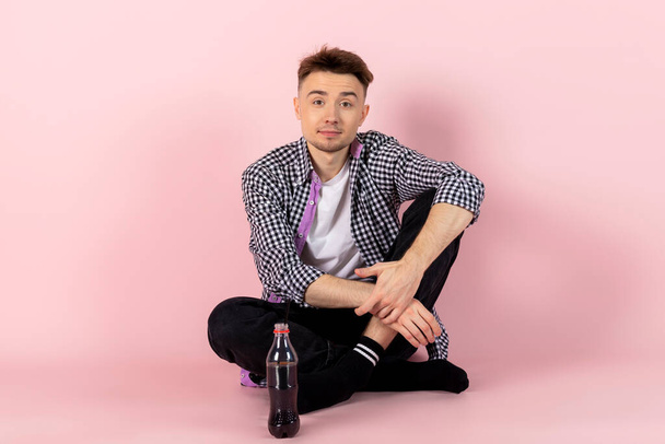 A happy young man sitting with a bottle of coke. fast food and sugar addiction. Isolated on pink background. - Fotoğraf, Görsel