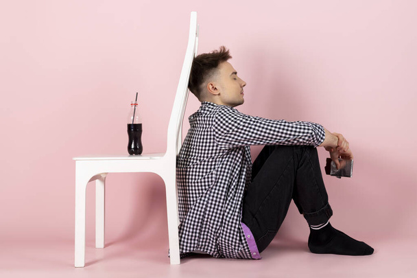 A young man holding a chocolate bar and turned his back to a bottle of coke or sweet soda. fast food and sugar addiction, unhealthy diet. Isolated on pink background. - Photo, Image
