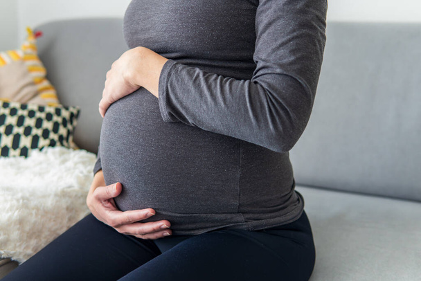 Pregnant woman sitting comfortably on the sofa and resting while putting her hands on her belly. Woman relaxing due to pregnancy pain and tiredness. Maternity, pregnancy concept. Front shot. - Foto, Bild
