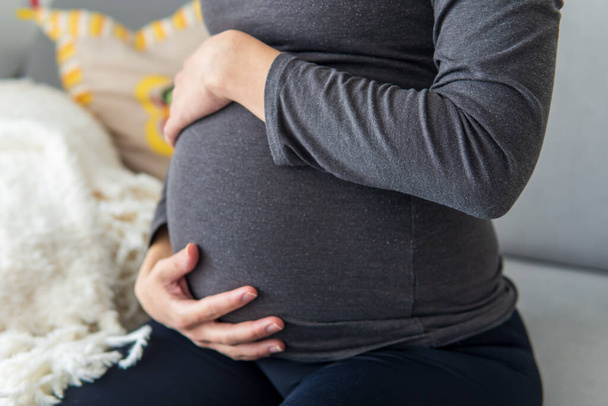 Pregnant woman sitting comfortably on the sofa and resting while putting her hands on her belly. Woman relaxing due to pregnancy pain and tiredness. Maternity, pregnancy concept. Front shot. - Foto, Bild