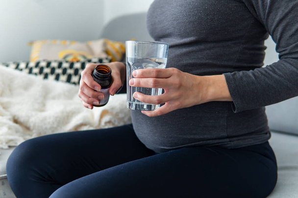 Беременная женщина принимает витамины и минералы из банки со стаканом воды, удобно сидя на диване. Беременность, здравоохранение и добавки концепция. Женщина принимает лекарства дома. - Фото, изображение