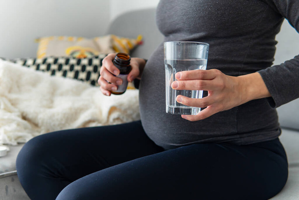 Беременная женщина принимает витамины и минералы из банки со стаканом воды, удобно сидя на диване. Беременность, здравоохранение и добавки концепция. Женщина принимает лекарства дома. - Фото, изображение