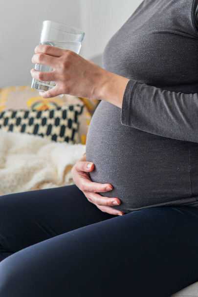 Schwangere, durstige Frau trinkt Wasser aus einem Glas, während sie gemütlich auf dem Sofa sitzt und ihren Bauch hält. Schwangerschaft, Gesundheitswesen und Hydratationskonzept. Gute Ernährung und Gewohnheiten während der Schwangerschaft. - Foto, Bild