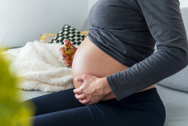  Ciężarna kobieta nakłada olejek ciążowy na jej pokazany brzuch, wygodnie odpoczywając na kanapie. Nawilżanie skóry pod kątem nawilżenia oraz zwalczanie rozstępów i suchości. Pojęcie macierzyństwa i ciąży - Zdjęcie, obraz