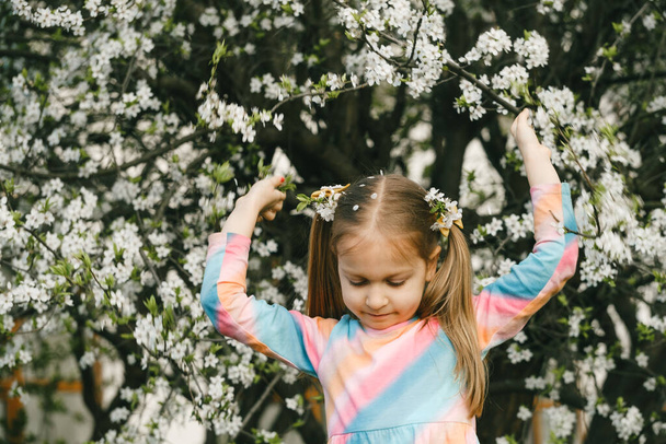 Nice little child in blooming garden exploring spring nature. Shaking tree making petals falling on her head. Fun in the garden. - Foto, imagen