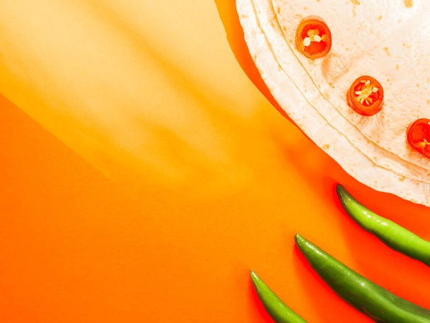 Three thin pita breads with sliced cherry tomatoes and three green chilli peppers on the right on an orange background with shadows and copy space on the left, flat lay close-up. Cinco de Mayo concept, minimalism, blanks. - Photo, Image