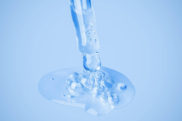 Transparentes Kosmetikgel fließt aus einer Pipette direkt in einem großen Klecks mit Blasen auf den Hintergrund. Auf blauem Hintergrund. Hyaluronsäure, Toner, Toner, Gel, Creme. Kosmetik. - Foto, Bild