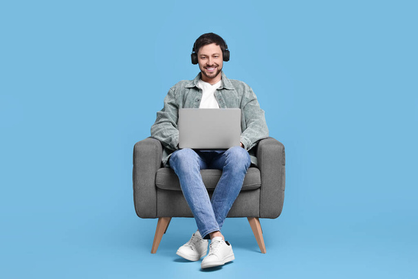 Happy man with laptop and headphones sitting in armchair on light blue background - Foto, Bild