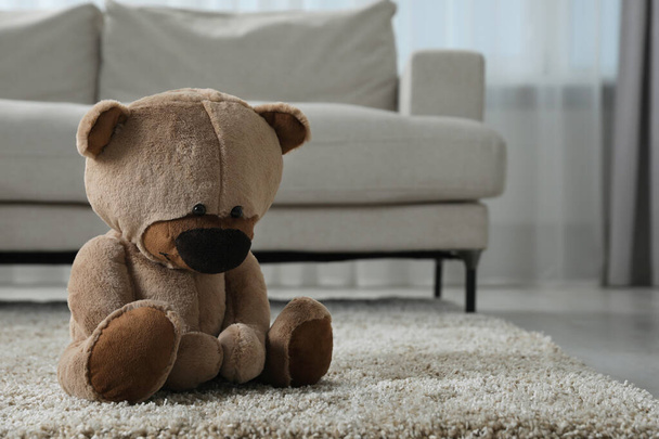 Cute lonely teddy bear on floor near sofa in room. Space for text - Foto, Imagem