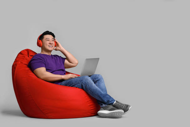 Smiling man with laptop and headphones sitting in beanbag chair against grey background - Fotografie, Obrázek