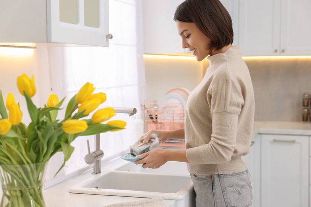 Happy young woman washing plate above sink in modern kitchen - Photo, Image