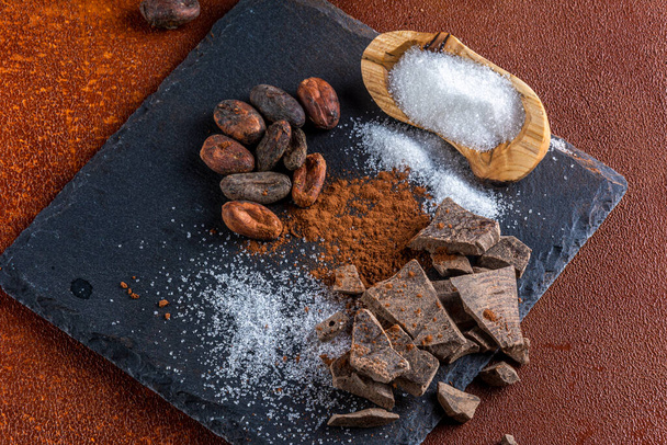 Pedazos de chocolate y sus ingredientes, sobre una losa de pizarra con elementos rústicos. - Foto, imagen