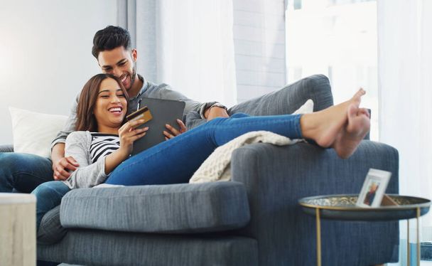 Im laughing but shes maxing my credit card. Full length shot of an affectionate young couple lounging on the sofa while using a tablet in their living room - Foto, imagen