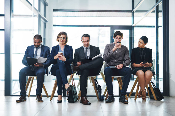 ここにいること自体が達成です。現代的なオフィスでのインタビューのために並んで座っているビジネスマンの多様なグループの完全な長さのショット - 写真・画像