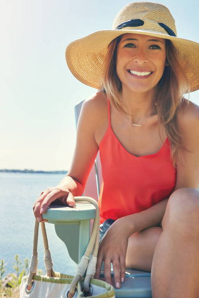 Making the best of the day doing nothing. Portrait of a cheerful young woman wearing a hat while being seated on a chair next to a lake outside in the sun - Photo, image
