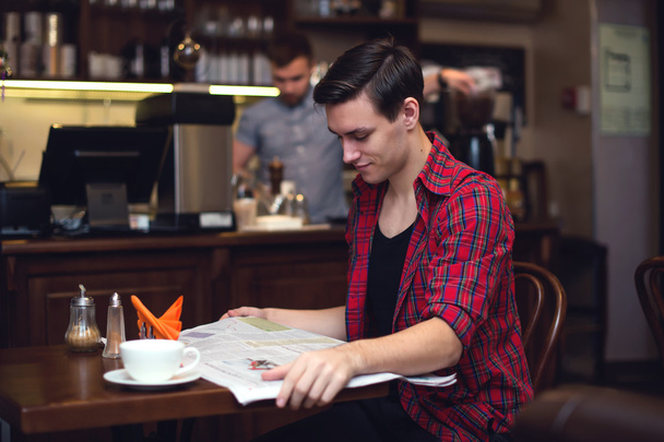Jeune étudiant attrayant boire du café et lire le journal dans le café avec barista sur le dos
 - Photo, image