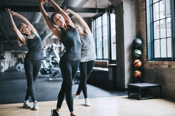 Stretch yourself. Full length shot of three attractive and athletic women stretching before their workout in the gym - 写真・画像