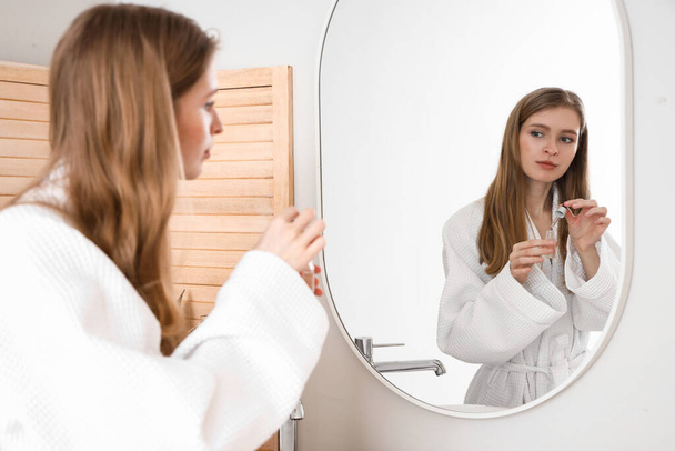 Νεαρή γυναίκα που χρησιμοποιεί ορό για την ανάπτυξη των μαλλιών κοντά στον καθρέφτη στο μπάνιο - Φωτογραφία, εικόνα