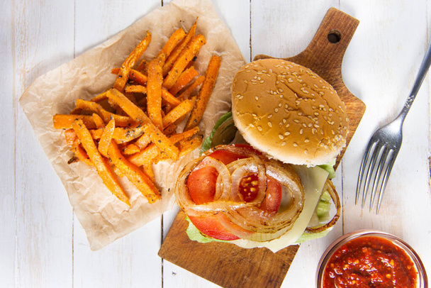 Homemade healthy burger variant, caramelized onions, fresh tomatoes, lettuce, homemade spice chilli ketchup, stocky chicken steak and homemade sweet potatoes fries on wooden white colored boards - Foto, Bild
