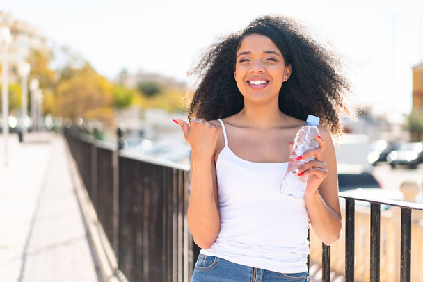 Fiatal afro-amerikai nő egy üveg vízzel a szabadban mutat oldalra, hogy bemutassa a terméket - Fotó, kép
