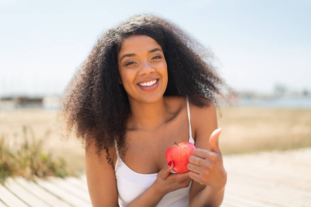Young African American woman with an apple at outdoors with thumbs up because something good has happened - Zdjęcie, obraz