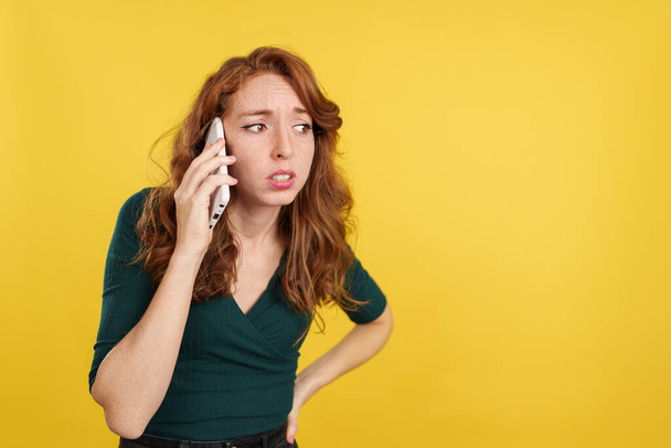 Bezorgde roodharige vrouw in gesprek met de mobiele telefoon in de studio met gele achtergrond - Foto, afbeelding