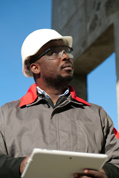 Αφροαμερικανός εργάτης με σκληρό καπέλο και προστατευτικά γυαλιά με tablet υπολογιστή στέκεται στο εργοτάξιο και κοιτάζει γύρω - Φωτογραφία, εικόνα