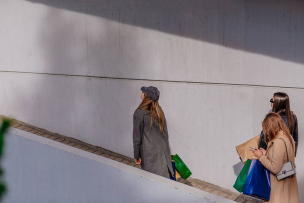Egy oldalnézeti fotó három elegáns lányról, amint besétálnak egy bevásárlóközpontba. Az egyik ellenőrzi a telefonját. - Fotó, kép