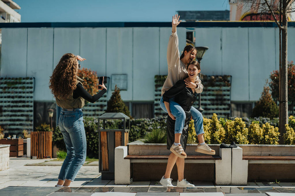 Curly haired girl taking a photo of her friends. Brunette girl giving a piggyback ride to her lovely friend - Фото, зображення