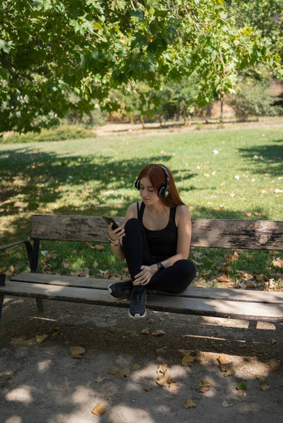 A young redhead girl, looking at the smartphone, with music headphones sitting on a park bench - Foto, afbeelding