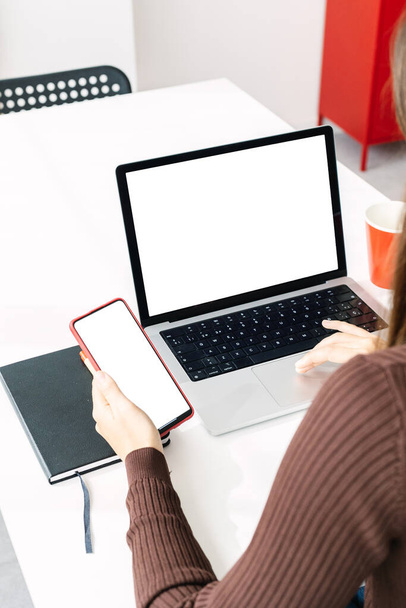 Imagem vertical da mulher segurando tela em branco simular telefone celular enquanto trabalhava no laptop em casa - Foto, Imagem