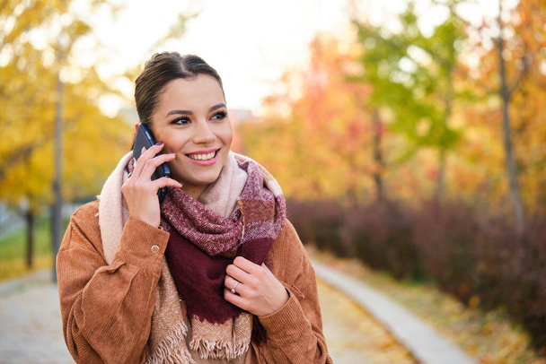 Mujer joven caucásica en una llamada telefónica sonriendo en otoño. - Foto, Imagen