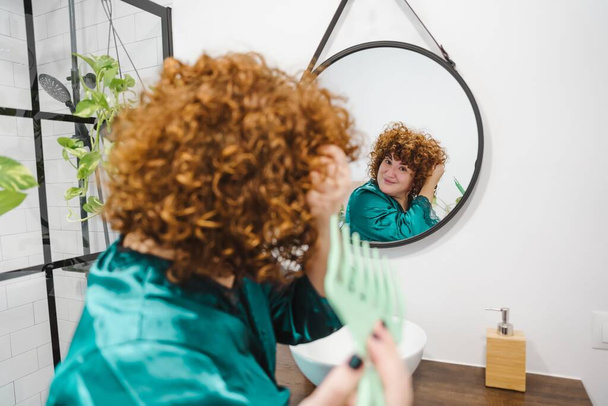 Een mollige vrouw met krullend gember haar borstelen haar haar in een badkamer spiegel - Foto, afbeelding