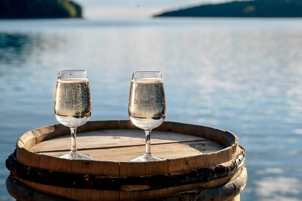 A closeup shot of two full wineglasses on the top of a wooden barrel against a blue sea - Foto, Imagen