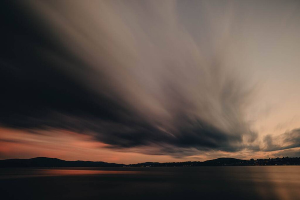 A long exposure shot of the gray clouds above a calm sea with the sunset in the background - Photo, Image