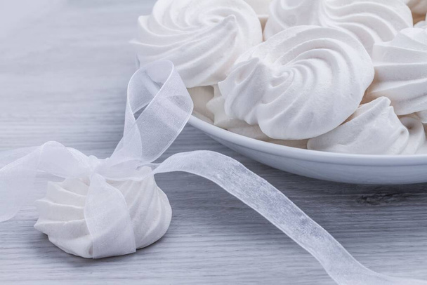 The meringue cookies on a white plate with another one with a bow on white wooden background. - Photo, Image